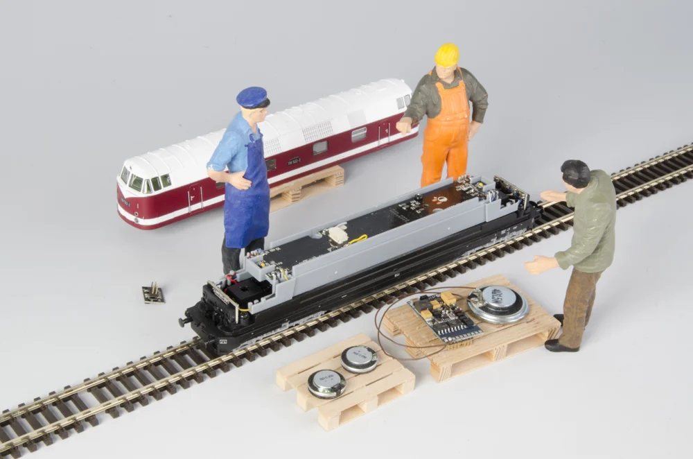 Geöffnete Modelllok mit Platine, Lautsprechern und Bauteilen, inszeniert mit Modellfiguren